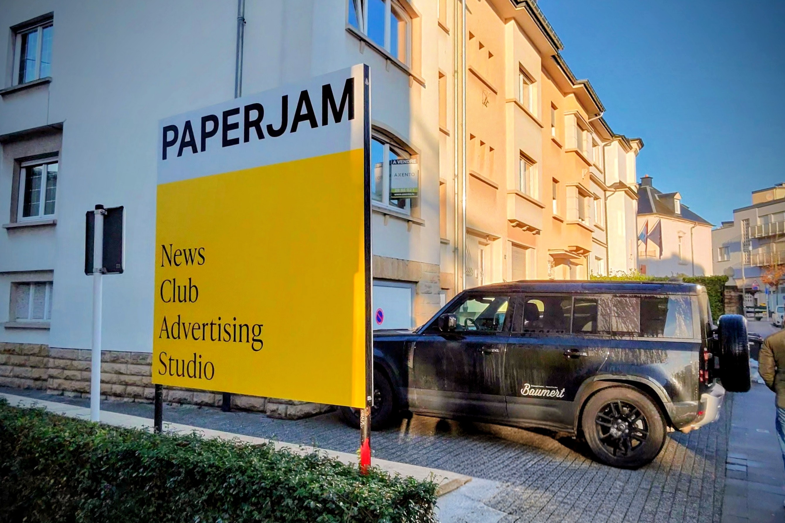Big yellow sign with the black & white logo of paperjam on top, with the mention of News, club, advertising and studio under. Street background with residential building and a 4x4 in parked behind the sign.